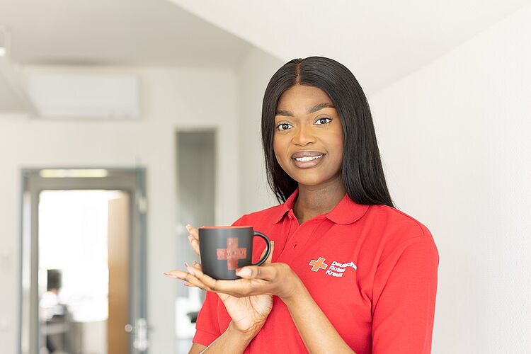 begegnungscafe01 Im Flur eines Bürogebäudes steht eine junge Frau mit langen, schwarzen Haaren. Sie trägt eine rotes Polo-Shirt mit Rotkreuz-Logo. In ihrer Hand hält sie eine schwarze Tasse mit Rotkreuz-Logo.